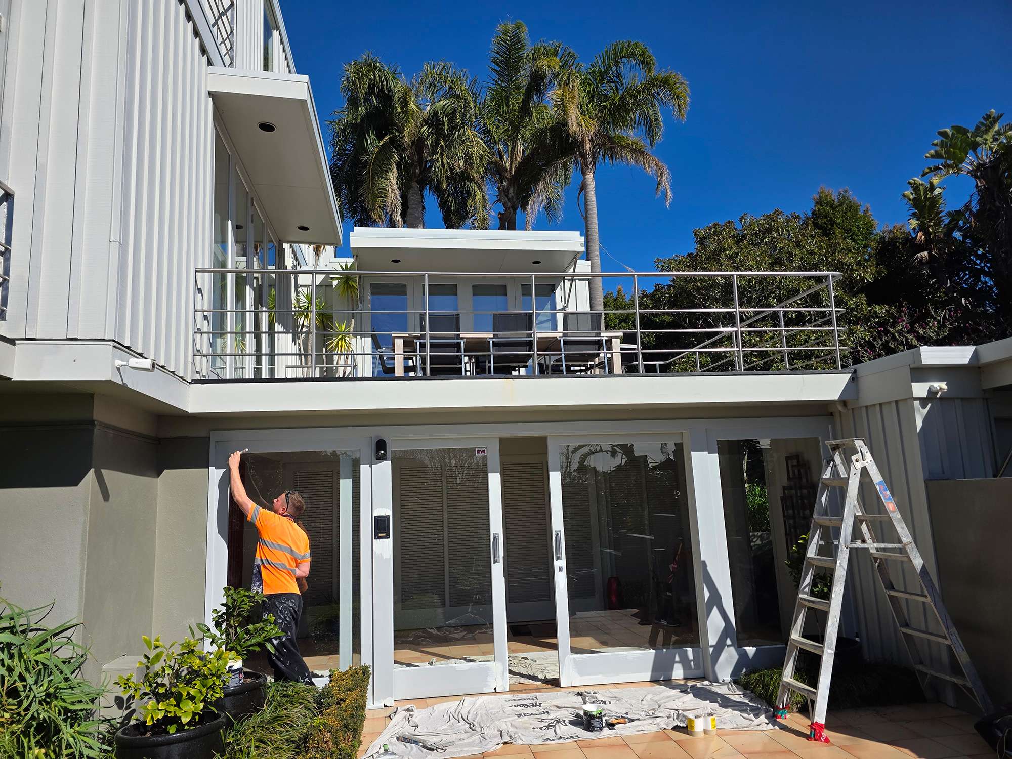 Window rot repair case study: Man painting exterior window frame, ladder nearby. Home exterior with balcony and palm trees.