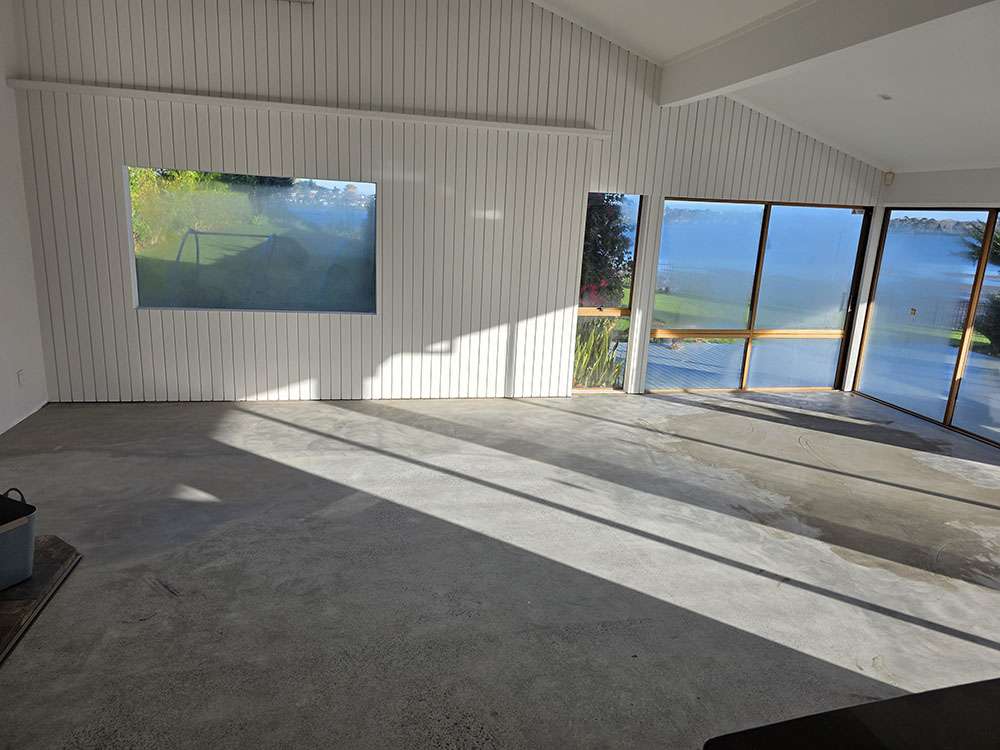 Modern living room with concrete flooring, large windows, and white wood paneling, showcasing natural light and minimalist design.