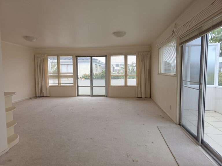 Empty light beige carpeted room needing paint and new flooring, with sliding doors and windows.