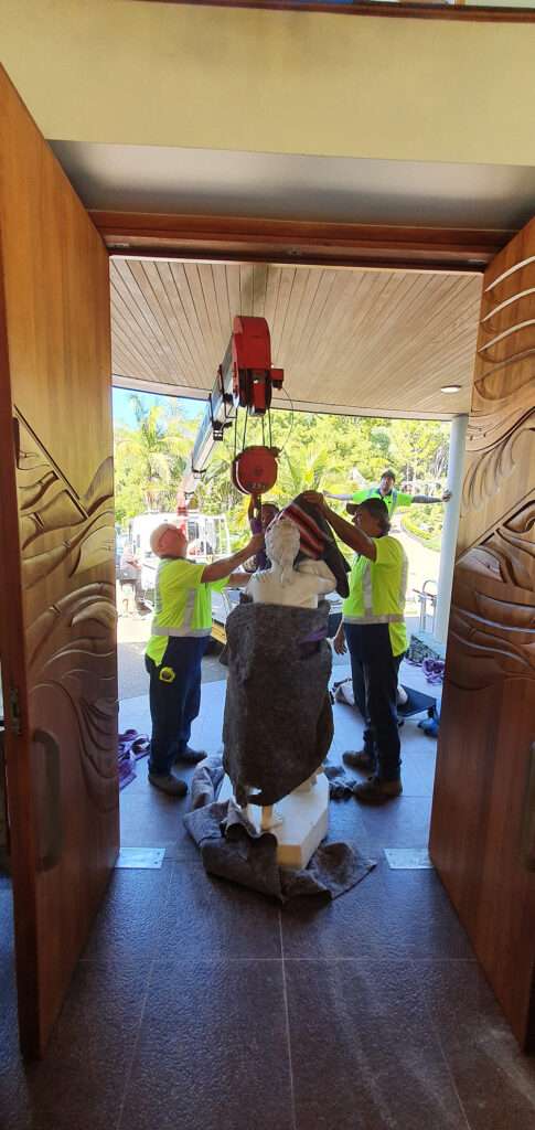 Hiab crane lifting statue off site. Workers in safety vests carefully maneuver the art piece during relocation.