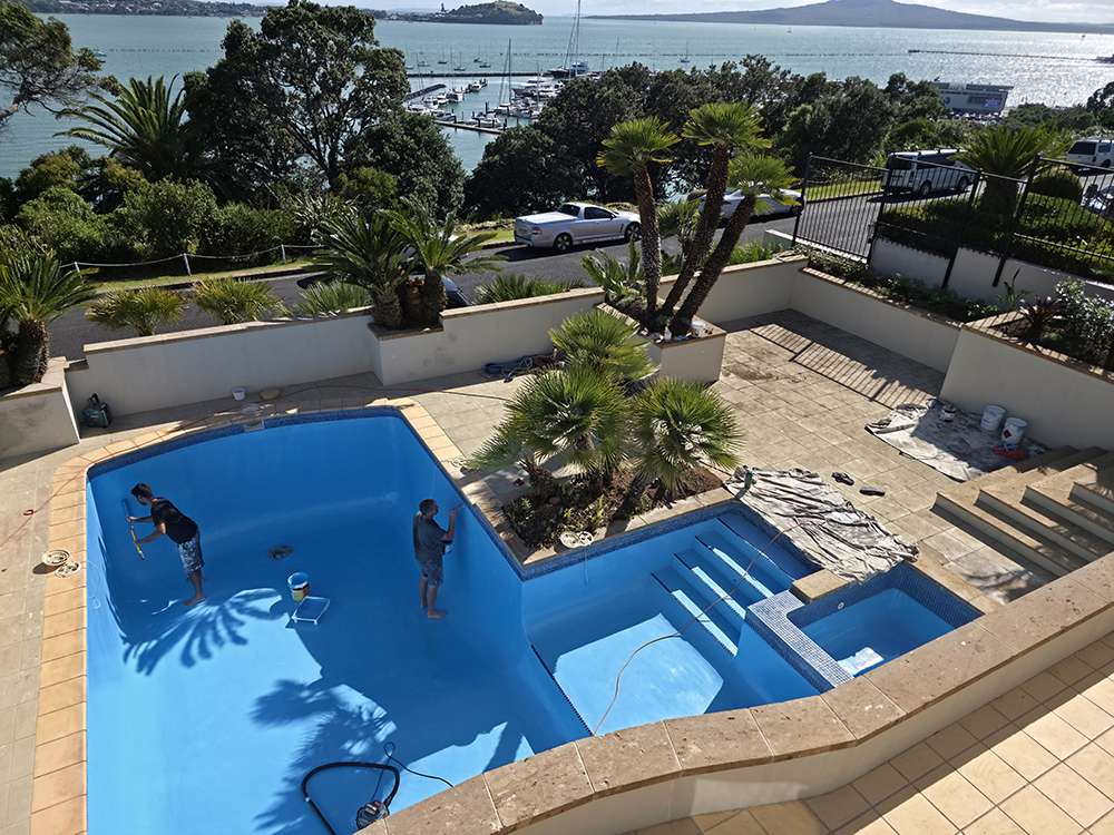 Pool painting in progress. Workers apply fresh blue paint. Pool pump repair, Auckland, New Zealand. Scenic ocean view.